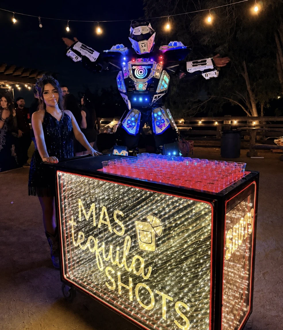 Quinceañera in a sparkling dress beside an LED robot and a lit tequila shot cart