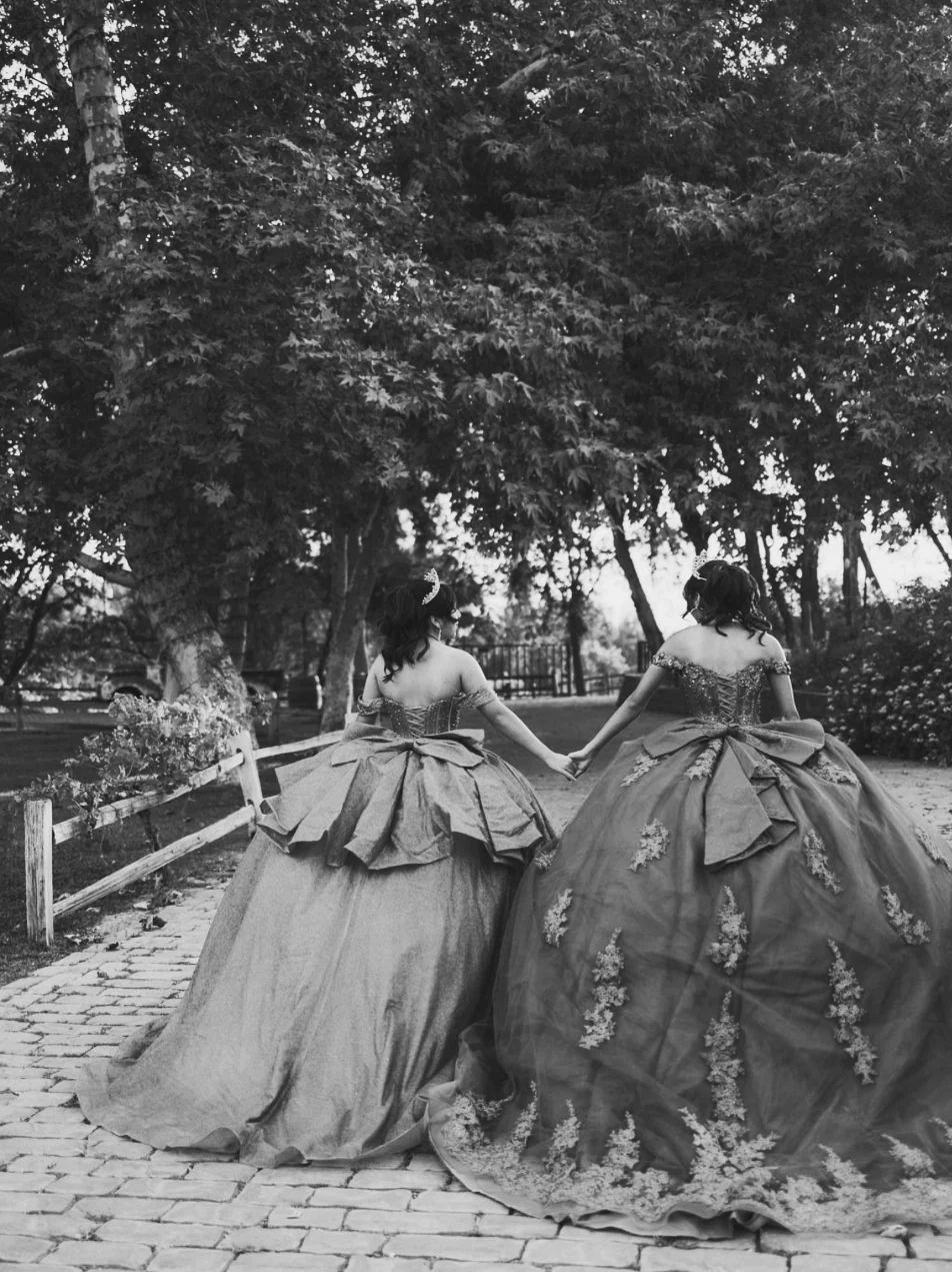 Two quinceañeras in ball gowns walking hand in hand down a garden path